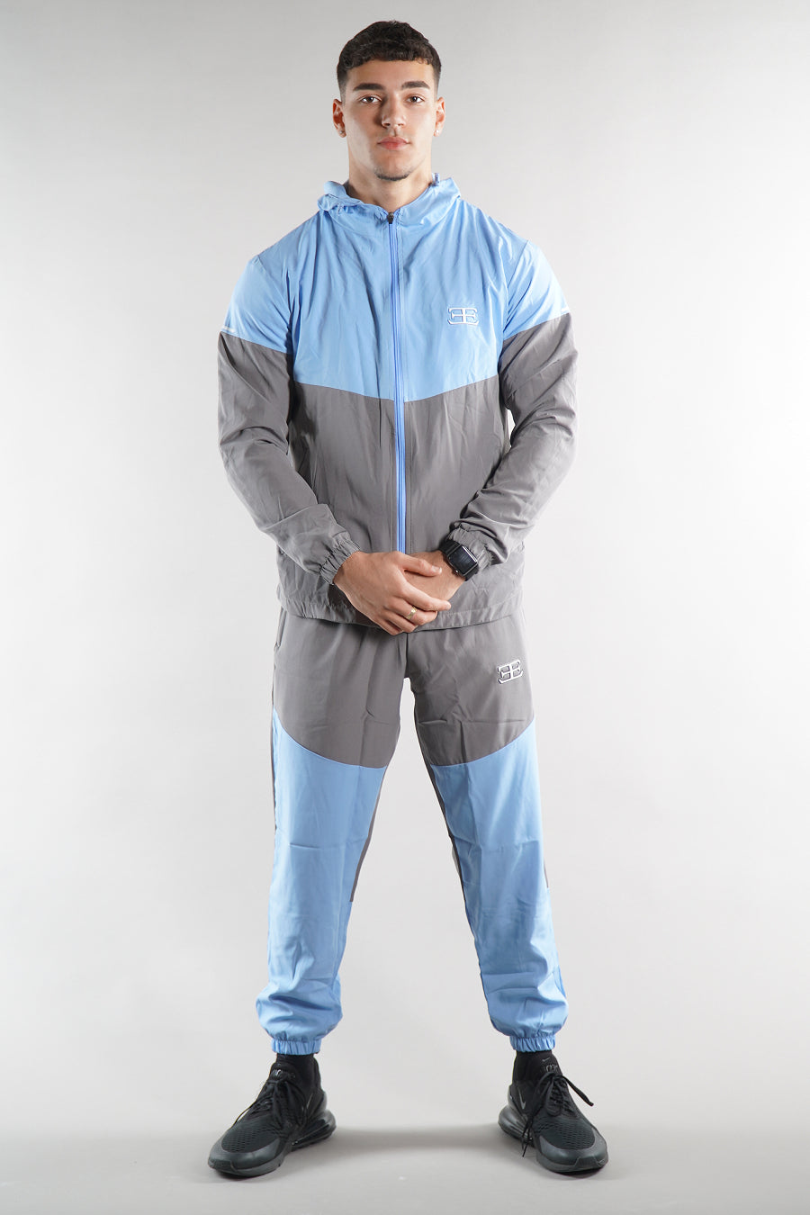 man wearing blue and grey elegante logo tracksuit jacket and joggers on white background